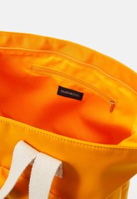 Żółta torba z tkaniny z białymi tekstylnymi uchwytami; posiada zamykaną na zamek kieszeń wewnętrzną oraz metkę wszytą w środku. Gładka tekstura na całej powierzchni.