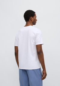 Maglietta bianca a maniche corte in cotone, con scollatura rotonda e texture liscia, vista da dietro, abbinata a pantaloni azzurri chiaro.