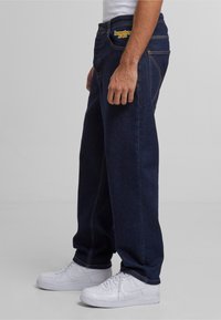 Jeans de mezclilla azul oscuro con un corte relajado, costuras visibles y logo en el bolsillo frontal. Llevados con zapatillas blancas.