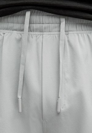 Lysegrå joggerbukser med elastisk talje, snøre og en glat tekstur. Har grå metaldele på snøreenderne.