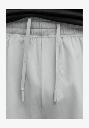 Svetlo sivi jogging hlače z elastiko v pasu, vrvico in gladko teksturo. Ima sivo strojno opremo na koncih vrvice.