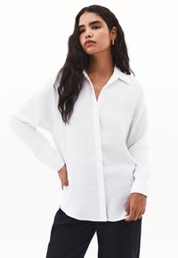 Camicia bianca oversize con bottoni in un tessuto testurizzato, caratterizzata da un colletto classico, maniche lunghe e una vestibilità comoda. Abbinata a pantaloni neri.