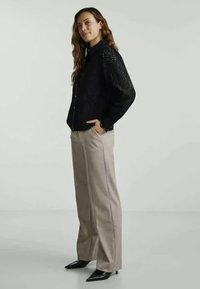 Musta nappikaulus paita, jossa on reikäkuviointia hihoissa, yhdistetty kevyisiin beigekeltaisiin leveälahkeisiin housuihin ja kärkipäisiin mustiin kenkiin.