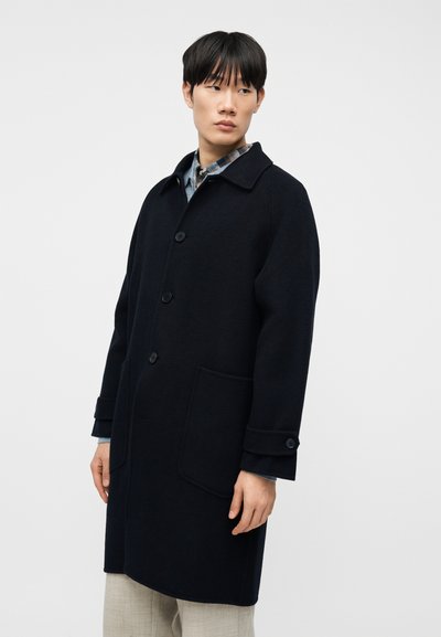 Νέος άνδρας που φοράει σκούρο ναυτικό κουμπωτό πανωφόρι πάνω από ένα πουκάμισο καρό και ανοιχτό γκρι παντελόνι, κοιτάζοντας ελαφρώς προς τα πλάγια με φόντο ένα απλό υπόβαθρο.