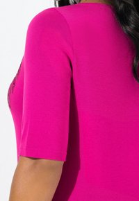 Top fucsia de manga corta con una textura suave, escote redondo y detalles decorativos en la zona de los hombros.