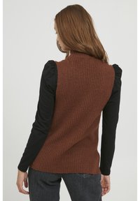Vesta din tricot de culoare maro, cu guler înalt, asortată cu un top negru cu mâneci lungi. Are un finisaj texturat și mici fante laterale.