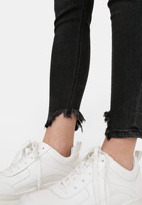 Pantalones vaqueros negros ajustados con dobladillo deshilachado, combinados con zapatillas blancas de suela plana y diseño con cordones. Material de mezclilla texturizado visible.