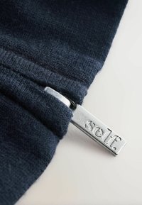 Tissu en polaire bleu marine à texture lisse, avec une tirette de fermeture éclair argentée marquée "self". Le tissu présente une légère peluche près de la fermeture éclair.