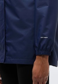 Veste imperméable bleu marine avec poignets élastiques, texture lisse et logo "DRYVENT" sur la manche. Gros plan sur le bras et le tissu.