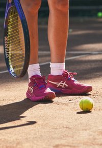 Vaaleanpunaiset tennarit, joissa on valkoisia yksityiskohtia ja violetteja elementtejä, valkoisten nilkkasukkojen kanssa, hiekka- kentällä tennispallon vieressä.