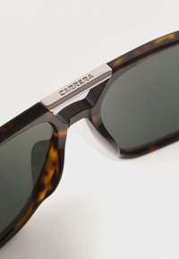 Tortoise shell zonnebril met donkere glazen. Heeft een zilveren metalen accent met "CARRERA" merknaam op de neusbrug. Gladde, glanzende oppervlakte.