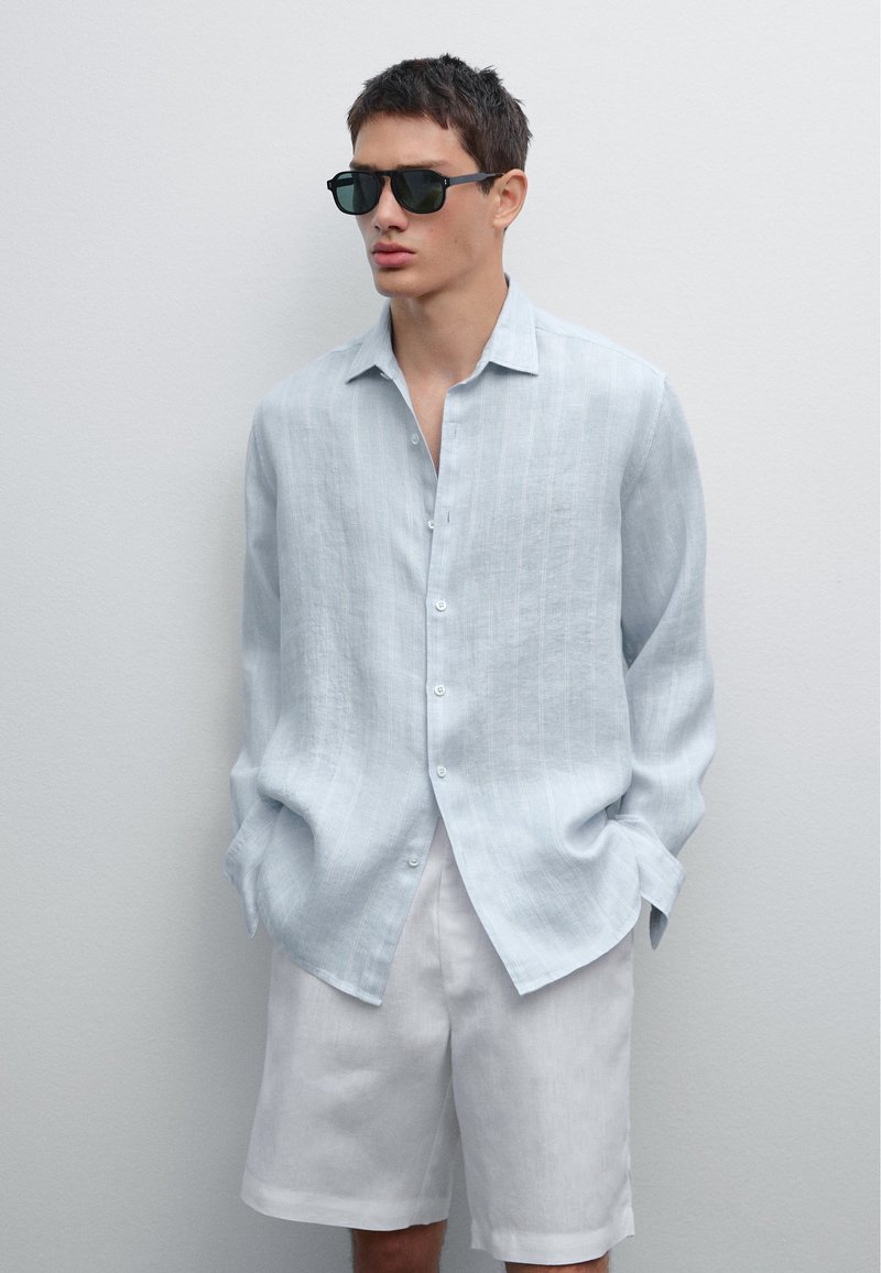 Camicia di lino azzurro chiaro con righe verticali, con bottoni, abbinata a pantaloni corti bianchi. Il modello indossa occhiali da sole neri. Taglio semplice e rilassato.