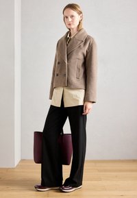 Woman standing indoors wearing a brown wool jacket, beige shirt, black wide-leg pants, burgundy shoes, and holding a large burgundy tote bag.