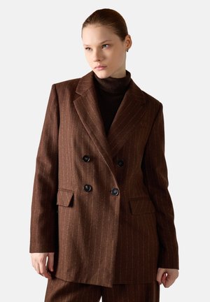 Vrouw draagt een bruin dubbelgeknoopt blazer met krijtstreep en bijpassende broek over een donkerbruine coltrui.