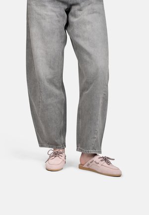 Onderbenen in losse grijze spijkerbroeken die lichtroze instap-mocassins dragen met bruine en witte veters op een lichte achtergrond.