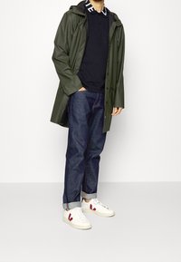 Zielony płaszcz przeciwdeszczowy z zapięciem z przodu, noszony na czarnej koszulce polo. W połączeniu z niebieskimi dżinsami i białymi trampkami z czerwonymi akcentami.