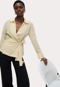 Camicia avvolgente a righe crema e beige, con scollatura a V profonda, maniche lunghe e dettaglio in vita annodato, abbinata a pantaloni larghi scuri.