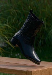 Black patent leather ankle boot with a crocodile skin texture, pointed toe, and low heel, displayed against a green background.