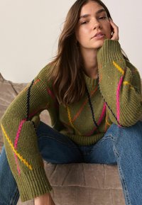 Knit sweater in olive green with multicolored diamond patterns, featuring ribbed cuffs and a relaxed fit. Paired with blue denim jeans.