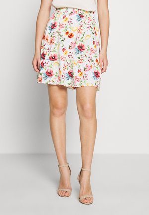 Vrouw draagt een witte bloemenrok met kleurrijke bloemen, gecombineerd met beige sandalen met hoge hakken, staand tegen een effen achtergrond.