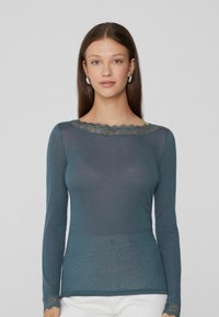 Langarmoberteil in tiefem Petrol mit Spitzenbesatz am Ausschnitt und an den Manschetten, mit einer taillierten Silhouette und leichtem, strukturiertem Stoff.