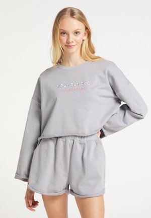 Giovane donna che indossa una felpa grigio chiaro con fiori di margherita e la scritta "Flower Girl", abbinata a pantaloncini con vita elastica, posando con la mano sul fianco.