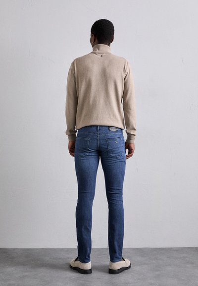 Pulover bej cu guler înalt, asortat cu blugi din denim albastri. Blugii au un design al buzunarului din spate și o nuanță decolorată.