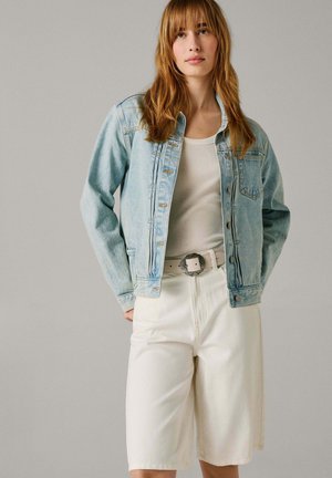 Persona joven con cabello largo castaño claro, que lleva una chaqueta vaquera azul claro, una camiseta blanca de canalé, pantalones cortos blancos de pierna ancha y un cinturón blanco con hebilla plateada.