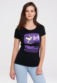 Black cotton t-shirt featuring a purple graphic of a cartoon bird wearing sunglasses, text reading "'69 WOODSTOCK." Fitted design, short sleeves.