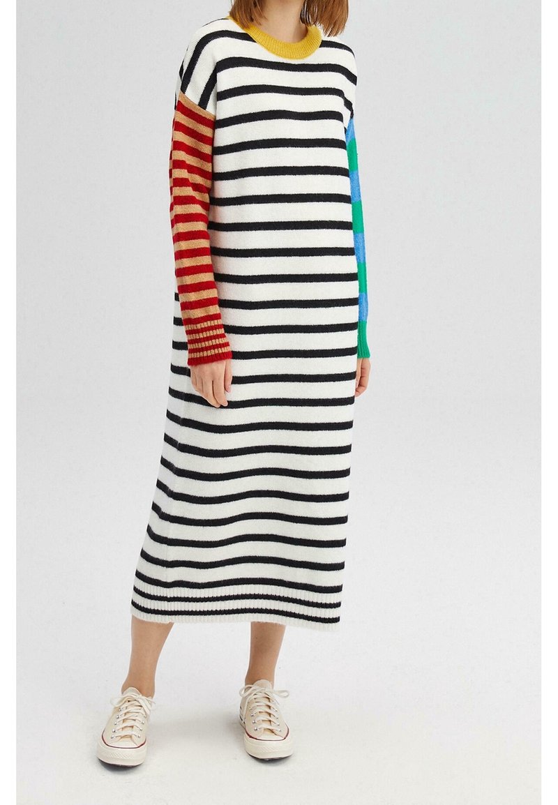 Personne portant une longue robe blanche avec des rayures horizontales noires et des manches dépareillées, associée à des baskets blanches à lacets.