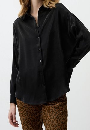 Vrouw draagt een losse zwarte satijnen blouse met knopen en een broek met luipaardprint, hand ontspannen langs haar zijde tegen een effen achtergrond.