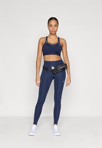 Tengerészkék sportmelltartó, hálós részletekkel, magas derekú tengerészkék leggings-szel párosítva, valamint egy fekete deréktáskával, amely vízpalack tartóval rendelkezik.