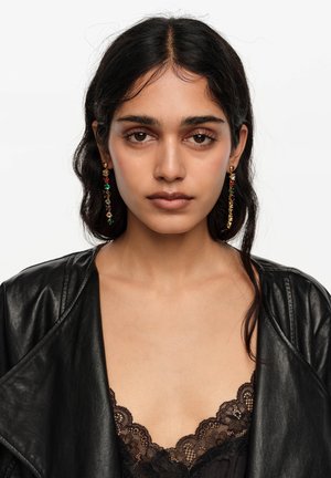 Joven con cabello largo y oscuro que lleva pendientes colgantes multicolores, blusa de encaje negra y chaqueta de cuero negra, mirando directamente al frente.