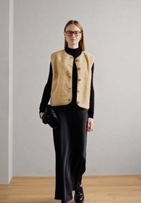 Beige texturerad väst med runda knappar över en svart polo och lång svart klänning. Modellen bär en svart kuvertväska. Trägolv, neutral bakgrund.