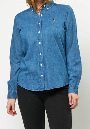 Kvinde iført en knaplukket blå denimskjorte med en lille broderet logo og sorte bukser, stående mod en ensfarvet baggrund.
