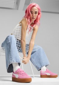 Sneakers di camoscio rosa con accenti bianchi e suola in gomma, abbinate a jeans in denim a gamba larga azzurri e calze bianche, su una sedia.