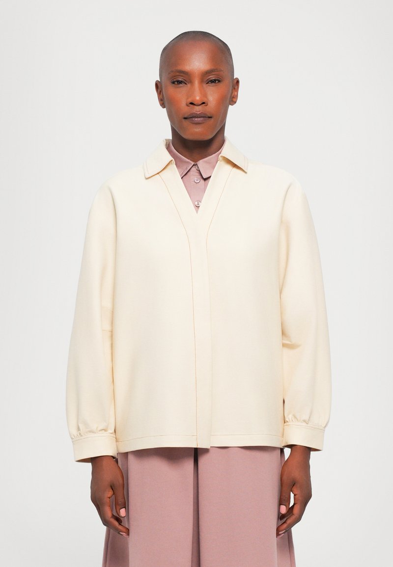 Crème-kleurige blouse met lange mouwen en een V-hals, voorzien van een gestructureerde kraag, elastische manchetten en contrasterende stikdetails.