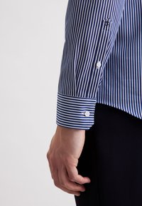 Camicetta con bottoni, a righe verticali blu e bianche, con polsini a righe contrastanti, bottoni bianchi e una texture liscia.