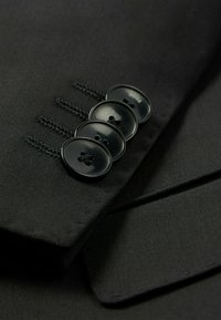 Il tessuto della giacca nera è caratterizzato da una vista ravvicinata di quattro bottoni neri con un logo sottile, cuciture dettagliate e una consistenza liscia.