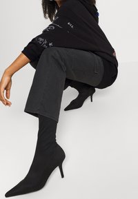 Persona agachada vistiendo un sudadera gráfica negra, pantalones anchos oscuros y botas de tacón de punta negra en un suelo blanco.