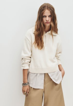 Crèmekleurige gebreide pullover met een ritssluiting bij de kraag, een ribgebreide onderkant, gedragen over een gestreepte overhemd met knopen en tankleurige wijde broek. Zilveren armbanden en ringen.