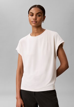 Vrouw met kort haar die een wit kort mouw blouse draagt en een zwarte broek, staand met één arm achter haar rug tegen een grijze achtergrond.