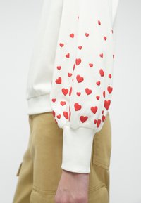 Baltā sporty kreklā ar sarkaniem sirdīm uz piedurknēm. Audums izskatās mīksts, ar ribota aproces detaļu un brīvu piegriezumu.