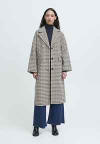 Donna che indossa un lungo cappotto a quadri beige e blu navy sopra un dolcevita blu navy, jeans a gamba larga blu e stivali neri alla caviglia, in piedi su uno sfondo neutro.