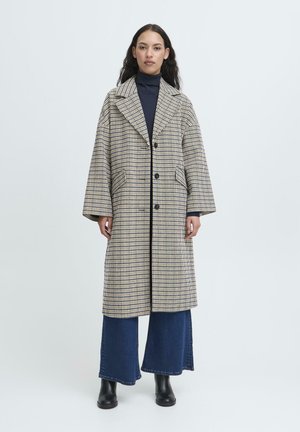 Donna che indossa un lungo cappotto a quadri beige e blu navy sopra un dolcevita blu navy, jeans a gamba larga blu e stivali neri alla caviglia, in piedi su uno sfondo neutro.