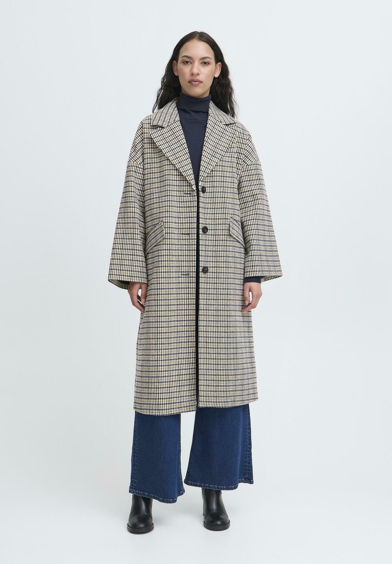 Donna che indossa un lungo cappotto a quadri beige e blu navy sopra un dolcevita blu navy, jeans a gamba larga blu e stivali neri alla caviglia, in piedi su uno sfondo neutro.