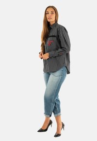 Donna con capelli lunghi che indossa una camicia a righe nere con dettagli rossi, jeans azzurri a risvolto e tacchi alti neri, in piedi su uno sfondo bianco.