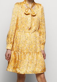 Robe fleurie jaune avec un décolleté à nouer, texture plissée, manches longues et ourlet à volants, présentant un motif délicat d'accents noirs et blancs.