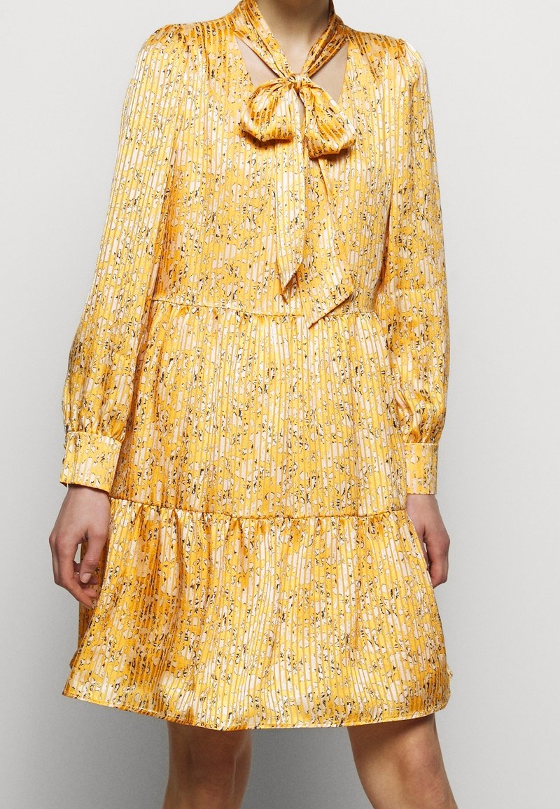 Robe fleurie jaune avec un décolleté à nouer, texture plissée, manches longues et ourlet à volants, présentant un motif délicat d'accents noirs et blancs.