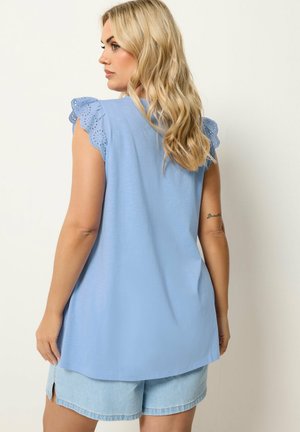 Blondynka ubrana w jasnoniebieski top bez rękawów z koronkowymi falbankami na ramionach i jasne szorty dżinsowe, zwrócona bokiem na jednolitym tle.